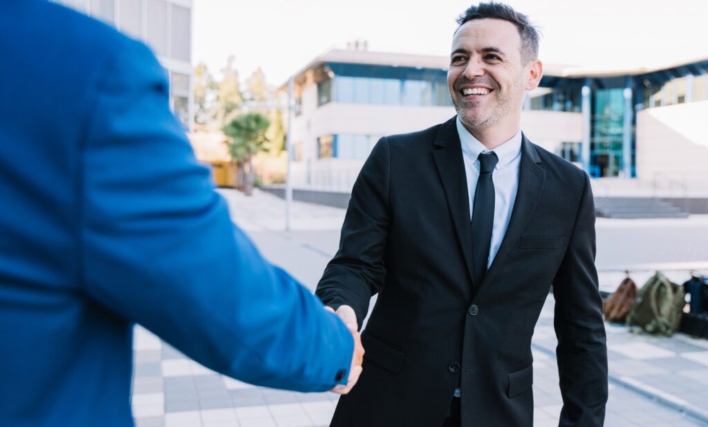 Smiling businessman in a black suit shakes hands with a colleague outdoors, modern office building in the background.