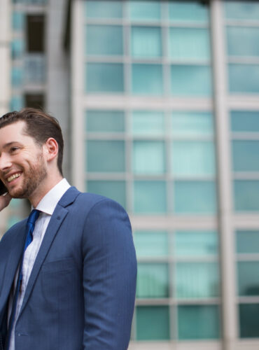 Smiling man in a blue suit talking on his smartphone outside a glass-fronted office building.