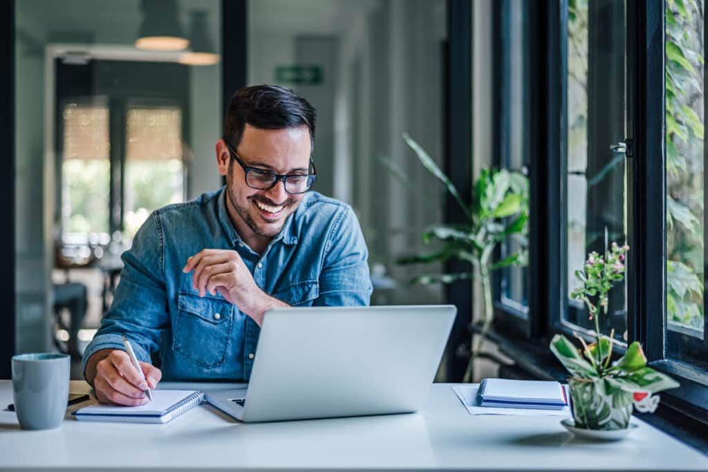 Smiling man in a denim shirt writes in a notebook while using a laptop at a bright desk by a window with plants nearby.