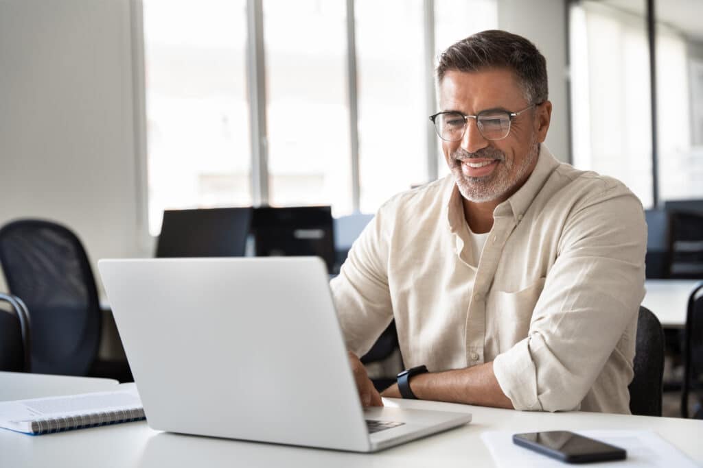 Smiling man with glasses working on a laptop at a bright office.